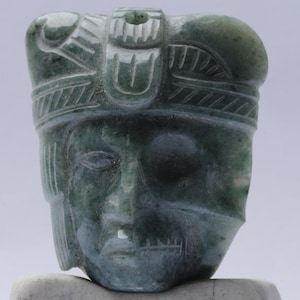 May include: A carved jade sculpture of a human head with a headdress. The sculpture is a dark green color with detailed facial features and headwear. The carving is set on a light gray base.