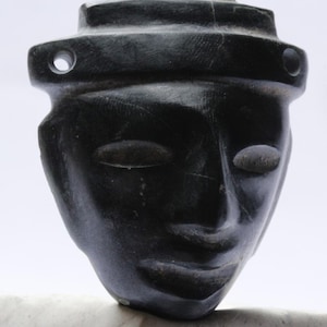 May include: Black stone sculpture of a face with closed eyes and a carved headdress. The sculpture is carved in a minimalist style.