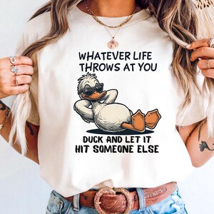 May include: Cream-colored t-shirt featuring a cartoon duck wearing sunglasses, reclining with its arms behind its head. The shirt has the text "WHATEVER LIFE THROWS AT YOU, DUCK AND LET IT HIT SOMEONE ELSE."