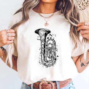 May include: Cream-colored t-shirt featuring a detailed black and white illustration of a tuba, surrounded by musical notes. The tuba design is centered on the shirt, creating a visually striking musical theme. The shirt is made of a soft material.