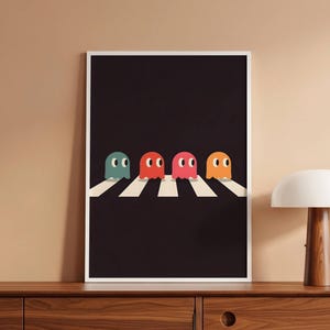 Op de afbeelding: Een ingelijste kunstdruk met vier kleurrijke spookkarakters die een wit zebrapad oversteken tegen een zwarte achtergrond. De geesten zijn in tinten van teal, rood, roze en oranje. De print wordt weergegeven op een houten kast.