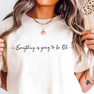 Peut inclure: T-shirt ivoire avec l'inscription "Everything is going to be Ok" en écriture noire. Le t-shirt est associé à un short en jean bleu clair et à une ceinture marron avec une boucle décorative. La tenue est accessoirisée de bijoux.