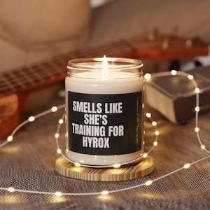 May include: A white candle with a black label that reads "Smells Like She's Training For Hyrox". The candle is lit and sitting on a wooden coaster.