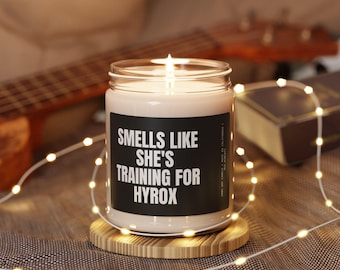 Pode incluir: Uma vela branca com um rótulo preto que diz "Smells Like She's Training For Hyrox". A vela está acesa e está sobre um suporte de madeira.