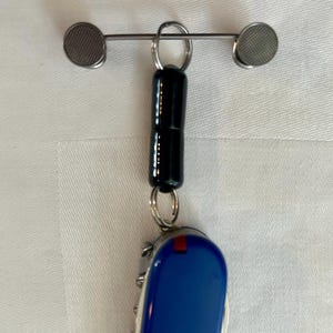 May include: A keychain featuring a black, cylindrical charm with a silver bar and two circular mesh accents. A blue and silver multi-tool knife hangs below the charm, with a red accent on the side.