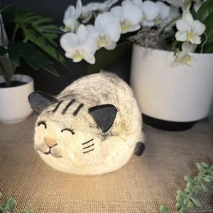 Puede incluir: Una luz nocturna de fieltro de gato blanco con ojos negros y bigotes. El gato está acostado con los ojos cerrados y tiene una expresión feliz en su rostro.