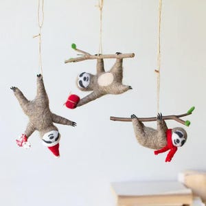 Può includere: Tre addobbi di bradipo in feltro appesi a uno spago. Ogni bradipo indossa un cappello di Babbo Natale rosso e tiene una piccola scatola regalo rossa. I bradipi sono appesi a dei rami.