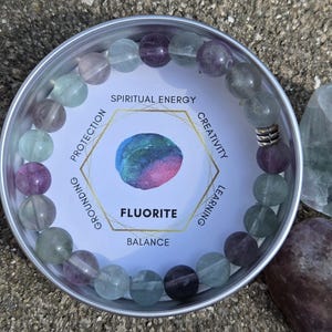 May include: A fluorite bracelet with multi-colored beads in a silver tin. The tin has a white label with the word "Fluorite" and the words "Spiritual Energy", "Creativity", "Learning", "Balance", "Grounding", and "Protection".