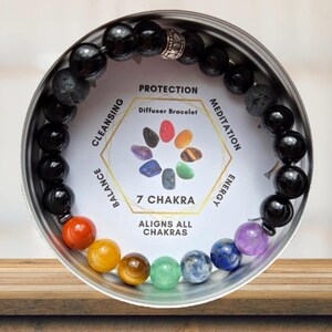 May include: A beaded bracelet with black and colorful stones, displayed in a silver tin. The bracelet features a variety of colored beads, including red, orange, yellow, green, blue, and purple. The tin has text that reads "7 Chakra" and "Aligns All Chakras."