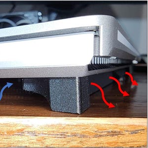 May include: A silver laptop computer on a black stand. The laptop has multiple ports and vents. Blue arrows indicate airflow direction from the front, and red arrows show airflow out the back. The laptop is on a wooden surface.