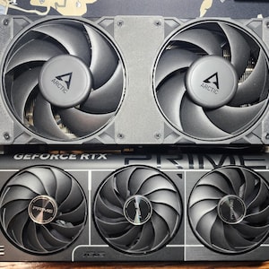 Könnte beinhalten: Zwei Computer-Grafikkarten werden gezeigt, eine über der anderen. Die obere Karte hat zwei große Lüfter mit dem "ARCTIC"-Logo. Die untere Karte hat drei Lüfter und den Text "GEFORCE RTX PRIME". Beide Karten sind grau und schwarz.