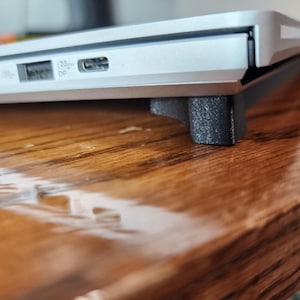 Könnte beinhalten: Ein silberner Laptop mit einem schwarzen Gummiständer auf einer Holzoberfläche. Der Laptop hat mehrere Anschlüsse an der Seite, darunter einen USB-C-Anschluss.