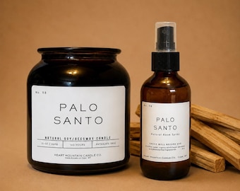 Puede incluir: Un frasco de vela de vidrio ámbar oscuro y un atomizador de vidrio ámbar a juego, ambos etiquetados Palo Santo No. 98 de Heart Mountain Candle Co. La vela pesa 369g y el spray 118ml. Varios palitos de madera clara están apilados detrás de ellos.