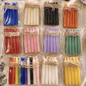 May include: Ten bundles of thin taper candles, each wrapped in clear plastic and tied with twine featuring a small metal charm. The sets include solid colors of blue, white, black, orange, red, pink, purple, green, yellow, and a multi-color pack.