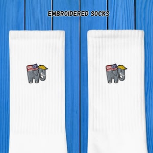 May include: White socks with an embroidered elephant wearing a yellow hat and a flag-themed bandana. The socks have the text "EMBROIDERED SOCKS" at the top. The socks are displayed against a blue wooden background.