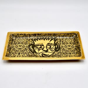 May include: A rectangular ceramic tray with a black and white graphic of a face on a yellow background. The face has a black outline and a black and white patterned background.
