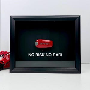 May include: A red Ferrari car key displayed in a black shadow box. The key is centered against a black background, with the text "NO RISK NO RARI" in white below. A red toy car is partially visible on the left.