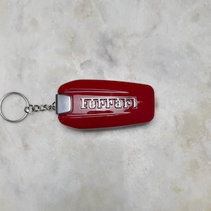 May include: A red Ferrari keyring with a silver metal accent and a silver key ring. The word "Ferrari" is displayed in silver lettering on the red keyring.