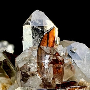 May include: Close-up of a cluster of quartz crystals with clear and brown hues. The crystals have sharp, geometric shapes and reflect light, creating a sparkling effect. The background is black, highlighting the crystals.
