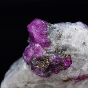 May include: Close-up of a raw ruby crystal cluster. The deep magenta-colored rubies are embedded in a matrix of white and gray stone, with flecks of gold-colored pyrite. The background is solid black.