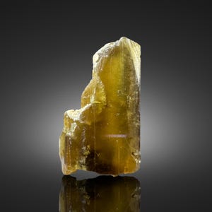 Könnte beinhalten: Ein durchscheinender, olivgelber Mineralkristall mit einer gestuften, vertikalen Struktur. Das Exemplar weist eine glatte, längliche Fläche auf der rechten Seite und eine gezackte, kristalline Textur auf der linken Seite auf.