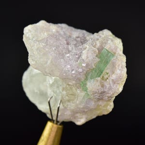 May include: A close-up of a raw mineral specimen featuring a light green tourmaline crystal embedded in a matrix of pale pink and white stone. The mineral is held by a brass and metal tool against a black background.