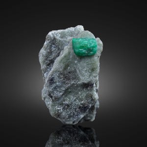 May include: A rough, gray rock formation with a vibrant, rectangular emerald crystal embedded in its surface. The emerald is a vivid green color, contrasting with the muted tones of the surrounding stone. The rock has a textured appearance.