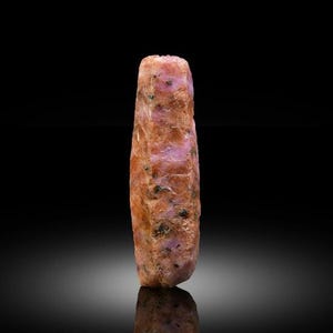 Può includere: Un cristallo di rubino grezzo e allungato con struttura esagonale, che mostra un mix di tonalità rosa opaco e arancione rossastro. La superficie presenta una consistenza naturale ruvida con piccole inclusioni minerali scure sparse.