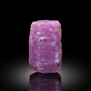 May include: A raw, uncut ruby gemstone with a vibrant pinkish-purple hue. The natural mineral displays a rough, textured surface and a rectangular shape. The gemstone is set against a black background, with its reflection visible below.