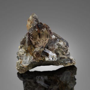 Puede incluir: Un grupo de cristales de cuarzo ahumado con tonos claros y marrones. Los cristales están parcialmente incrustados en una matriz de roca gris y marrón. La forma general es irregular, con puntas de cristal afiladas y una textura rugosa.