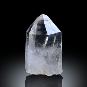 Op de afbeelding: Een heldere bergkristal punt met een gefacetteerde top en een ruwe, natuurlijke basis. Het kristal is transparant en laat licht door, tegen een zwarte achtergrond. De vorm van het kristal is een zeszijdig prisma, typisch voor kwarts.