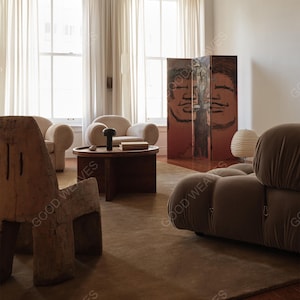 Könnte beinhalten: Ein Wohnzimmer mit einem braunen Sofa, zwei beigen Sesseln und einem Holzstuhl. Ein runder Couchtisch aus dunklem Holz mit einer schwarzen Lampe und Büchern. Ein dekorativer Raumteiler mit bemalten Gesichtern befindet sich im Hintergrund. Der Raum hat eine neutrale Farbpalette.