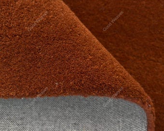May include: A solid rust-colored rug with a dense, short-pile texture folded over to reveal a light gray woven backing. The surface features a repeating watermark text that reads Good Weaves across the fabric in a diagonal orientation.