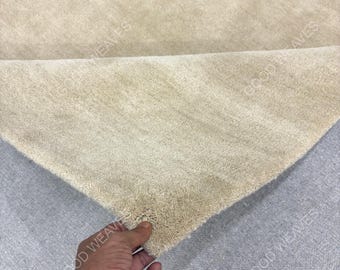 May include: A beige area rug with a thick, soft pile texture. The rug is folded over to reveal the dense, light-colored fibers and the woven backing. The text GOOD WEAVES is visible as a watermark across the surface of the rug.
