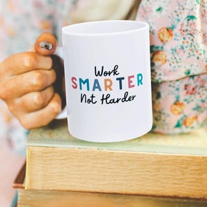 May include: White ceramic mug with the text "Work SMARTER Not Harder" in colorful letters. The mug is held by a person's hand and is sitting on top of a stack of books.