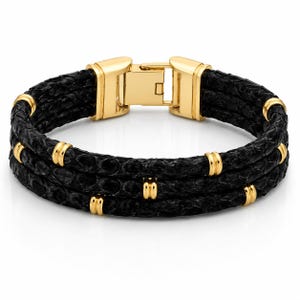 May include: A black textured leather bracelet featuring three parallel strands held together by polished gold-tone metal accents and a matching gold-tone rectangular box clasp closure. The design incorporates small gold-tone rings spaced along the strands.