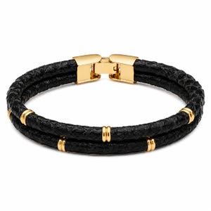 May include: A double-strand black textured bracelet featuring a polished gold-tone metal clasp and four small gold-tone metal rings spaced evenly along the two black bands, creating a layered look with a secure locking mechanism.