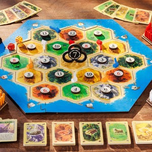 May include: A board game with a blue and tan hexagonal game board, surrounded by cards, wooden pieces, and dice. The game board features numbered spaces and colourful terrain. The game is called "The Semak".