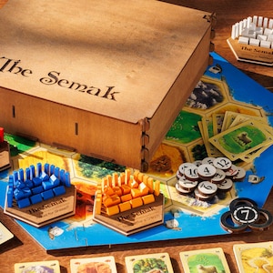 Può includere: Una scatola di legno con l'iscrizione "The Semak" è aperta, rivelando un gioco da tavolo con pezzi colorati e un tabellone di gioco blu. Il tabellone di gioco ha una griglia esagonale con numeri e simboli. Ci sono anche carte e gettoni sul tabellone.