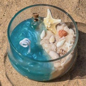 May include: A round glass bowl candle featuring a split design with translucent blue gel wax on one side and textured beige wax on the other. The beige section is decorated with a small corked glass bottle and various seashells and a starfish.