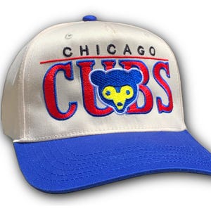 May include: Cream baseball cap with a blue peak. The cap displays the word "CHICAGO" above "CUBS" in red, with a blue bear logo. A classic sports accessory.