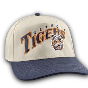 May include: A beige baseball cap with a navy blue peak. The cap displays the text "DETROIT TIGERS" in navy blue with orange accents, and a tiger logo patch. The cap has a navy blue button on top.