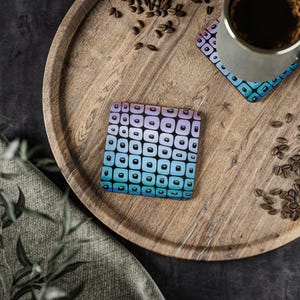 May include: Square coaster with a gradient design of blue, purple, and black squares. The coaster is on a wooden tray with coffee beans and a mug. The coaster has a matching design to the coaster under the mug.