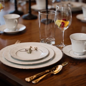 May include: A table setting with white plates, a white teacup and saucer, a glass of fruit punch, and gold silverware. The table is made of wood and has a dark brown finish.