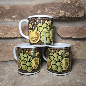 May include: Three white ceramic mugs with a brown border and a fruit design featuring green grapes and a yellow apple. The mugs are stacked on a stone surface against a brick background.