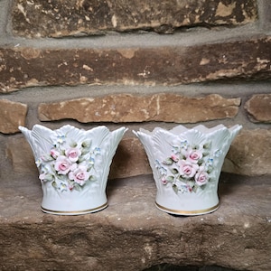 May include: Two white porcelain vases with a scalloped rim and gold trim. Each vase is decorated with pink roses and blue flowers. The vases are displayed on a stone surface against a brick background.