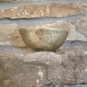 May include: A small, round, brass bowl with an etched design. The bowl is sitting on a rough, textured stone surface.