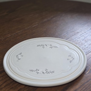 May include: Off-white, oval-shaped ceramic serving platter with a floral design. The platter has a scalloped edge and a raised rim. The floral design is in a muted gray color and is placed in the corners of the platter.