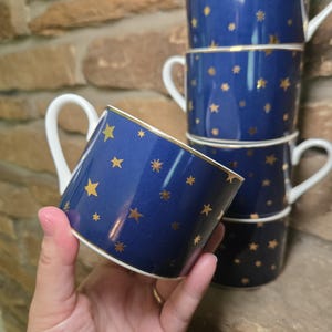 May include: A stack of navy blue ceramic mugs with gold star patterns. Each mug has a white handle and a gold rim. The mugs are stacked on top of each other, with one mug being held in a hand.