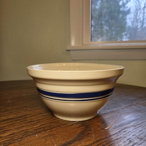May include: Cream-colored ceramic mixing bowl with two blue stripes. The bowl has a wide rim and a rounded shape. A classic kitchenware item, perfect for baking and food preparation.